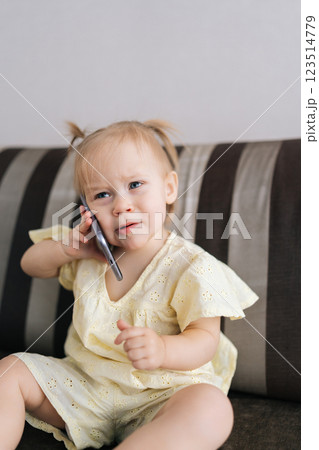 Vertical portrait of serious funny toddler girl listening intently to smartphone, pretending to make call while sitting on couch. Cheerful child expressing playful communication skills at home. 123514779