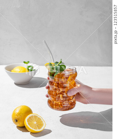 Hand holding glass of iced tea with lemon, ice and mint on light background with shadow and fruits. 123515857