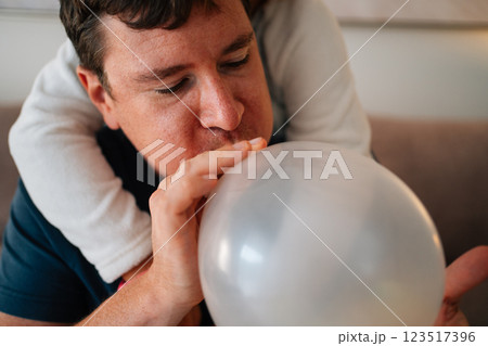 Caucasian man blowing balloon with child's arm wrapped around him indoors 123517396