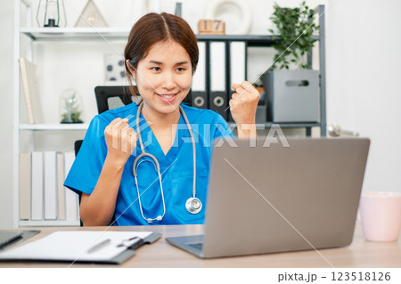 Happy nurse in blue scrubs celebrates success while using a laptop in a modern medical office setting. 123518126