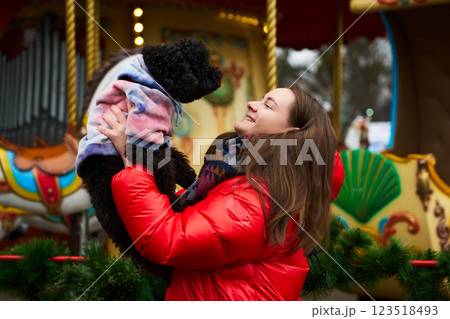 Woman joyfully lifts her dog in front of a colorful carousel during winter festivities in a park 123518493