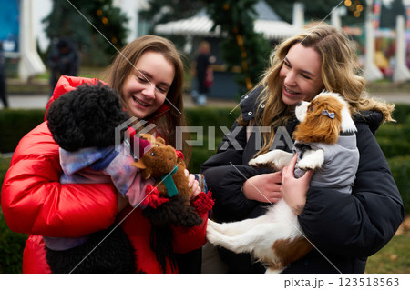 Two young women enjoy winter festivities while holding their adorable dogs in a park decorated for the holidays Two young women enjoy winter festivities while holding their adorable dogs in a park decorated for the holidays 123518563