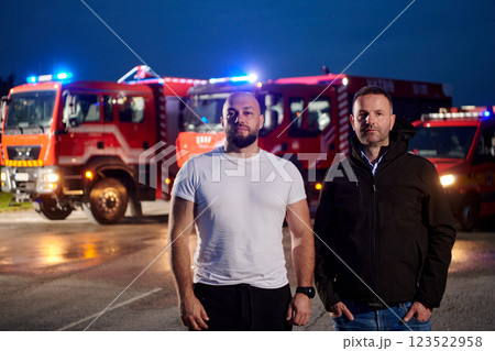 Group of firefighters, dressed in civilian clothing, stand in front of fire trucks during the night, showcasing a moment of camaraderie and unity among the team as they reflect on their duties and the 123522958
