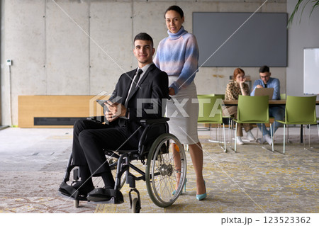 In a professional office setting, a businesswoman helping her director, who is sitting in a wheelchair and using a tablet, while their colleagues collaborate in the background. 123523362