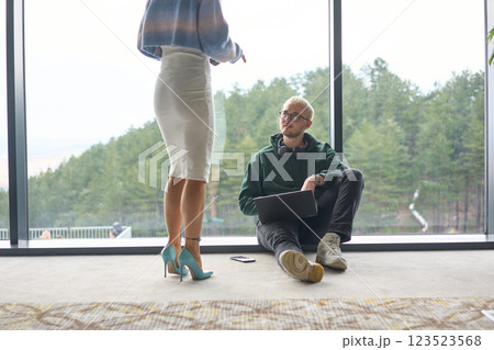 A blonde man sitting casually on the floor of a modern office, leaning against a window as he uses his laptop, with his attractive female colleague standing nearby 123523568