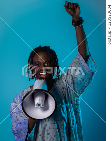 In a powerful and symbolic image, an African American man wears traditional clothing, passionately wields a megaphone against a striking blue background, with his hand raised in the air symbolizing 123523604