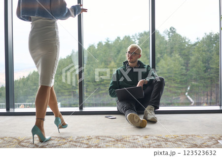A blonde man sitting casually on the floor of a modern office, leaning against a window as he uses his laptop, with his attractive female colleague standing nearby 123523632