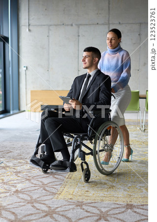In a professional office setting, a businesswoman helping her director, who is sitting in a wheelchair and using a tablet, while their colleagues collaborate in the background. 123523761
