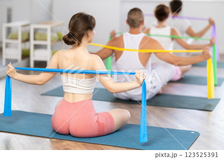 Back view of slim flexible athletic young woman sitting on sports mat practicing pilates during group exercises with elastic band in sports club Back view of slim flexible athletic young woman sitting on sports mat practicing pilates during group exercises with elastic band in sports club 123525391