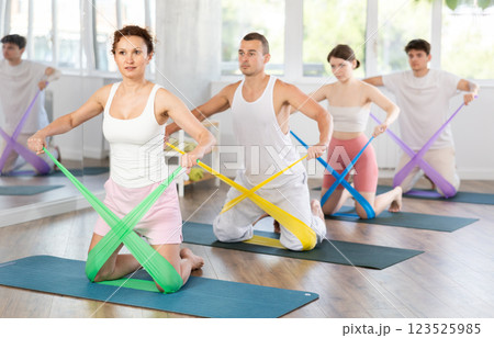 Sporty pretty young woman kneeling on mat and athletic women and men exercising with resistance bands in gym during group pilates class 123525985