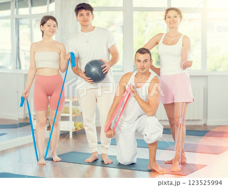 Young men and women posing together in training room holding resistance bands and pilates ball Young men and women posing together in training room holding resistance bands and pilates ball 123525994