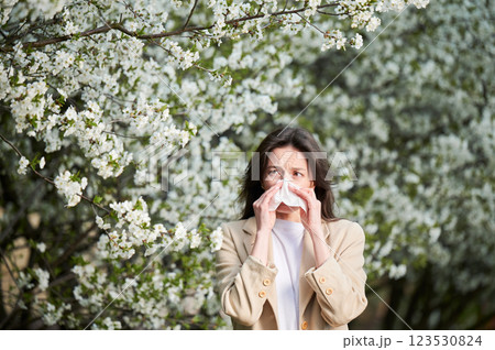 Woman allergic suffering from seasonal allergy at spring in blossoming garden at springtime. Young woman sneezing and blowing nose with nasal handkerchief in front of blooming tree. Allergy concept. 123530824