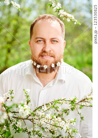 Man allergic enjoying after treatment from seasonal allergy at spring. Portrait of happy man with small white flowers dotted in beard, smiling in front of blossom tree at springtime. 123530825