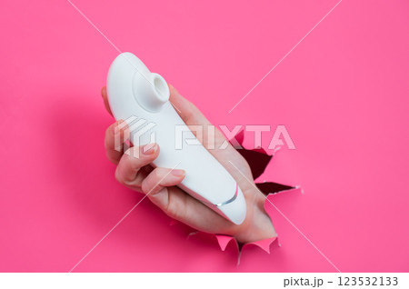 Female hand sticks out white vacuum clitoral vibrator through pink paper background. Female hand sticks out white vacuum clitoral vibrator through pink paper background. 123532133