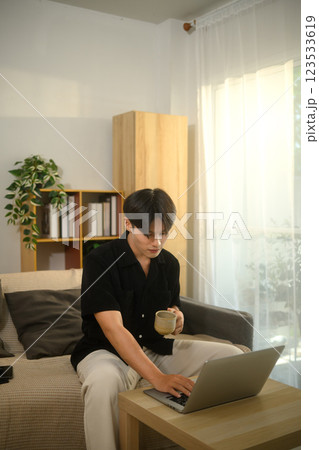 Young asian man holding a coffee cup and working on his laptop in a living room Young asian man holding a coffee cup and working on his laptop in a living room 123533619