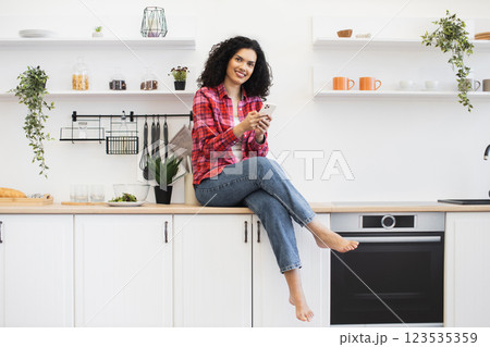 African American woman sitting on kitchen counter, wearing casual attire, using smartphone, smiling, modern kitchen decor. 123535359