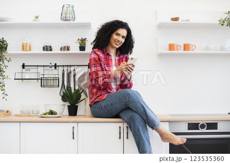 African American woman wearing casual outfit sits on kitchen counter holding smartphone smiling enjoying leisure time. 123535360