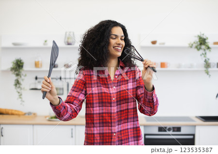 Young African woman, wearing vibrant plaid shirt, standing in kitchen, holding utensils, smiling energetically, expressing creativity and joy. 123535387
