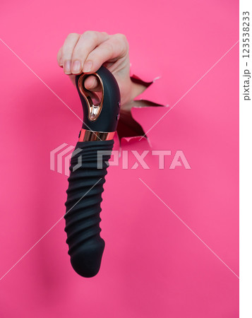 Female hand sticking out of pink paper background with black textured vibrator. Vertical photo. Female hand sticking out of pink paper background with black textured vibrator. Vertical photo. 123538233