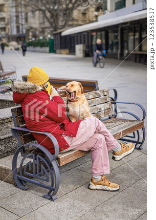 Person in red coat and yellow hat interacts with a playful dog on bench in a lively city square enjoying together and talking on phone. Cheerful atmosphere, modern technology Person in red coat and yellow hat interacts with a playful dog on bench in a lively city square enjoying together and talking on phone. Cheerful atmosphere, modern technology 123538517