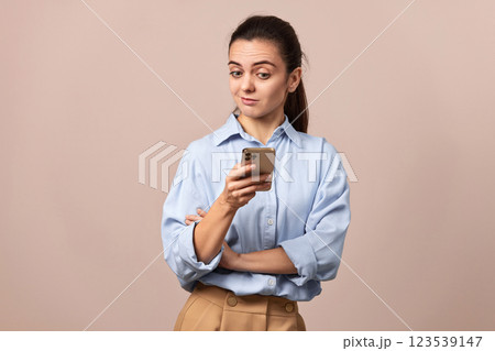 skeptical woman in blue shirt holding mobile phone looking at smartphone on beige background. 123539147