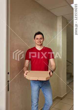 smiling delivery man in red t-shirt giving food order, holding pizza box standing at doorway smiling delivery man in red t-shirt giving food order, holding pizza box standing at doorway 123539285