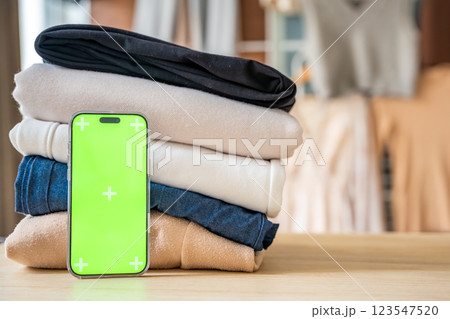 Stack of clothing prepared for resale with a smartphone displaying green screen for mockup use. Mobile apps simplify second-hand fashion, sustainable shopping, and conscious clothing redistribution 123547520