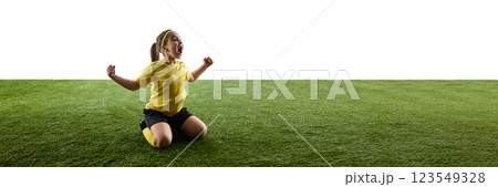 Emotional girl, soccer player in yellow jersey and black shorts sitting on grassy field, raising hands in victory pose against white background 123549328