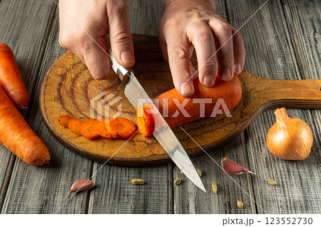 Hands skillfully slice a bright orange carrot on a wooden board, with garlic cloves and an onion nearby, showcasing a homely and engaging cooking atmosphere Hands skillfully slice a bright orange carrot on a wooden board, with garlic cloves and an onion nearby, showcasing a homely and engaging cooking atmosphere 123552730