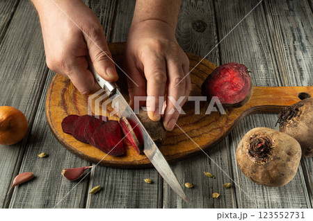 A pair of hands skillfully slices fresh beets on a wooden cutting board. Surrounding ingredients include garlic cloves and an orange, suggesting a vibrant culinary preparation A pair of hands skillfully slices fresh beets on a wooden cutting board. Surrounding ingredients include garlic cloves and an orange, suggesting a vibrant culinary preparation 123552731