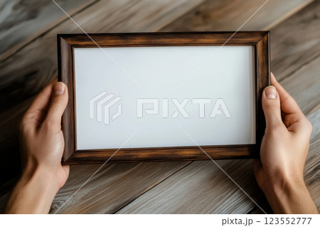 Empty frame mockup held by hands showcasing a blank picture frame on a wooden surface with natural tones and textures perfect for creative design purposes 123552777