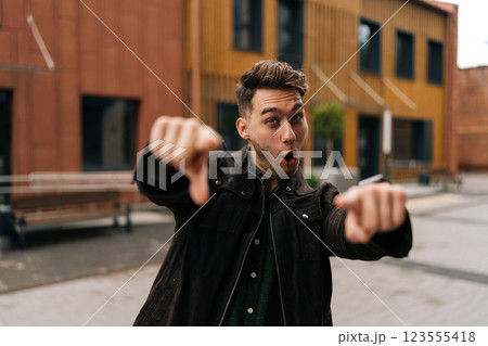 Waist up portrait of positive young man pointing fingers at camera with funny expression standing in city setting. Handsome happy male wearing casual clothes gesturing outside looking at camera. Waist up portrait of positive young man pointing fingers at camera with funny expression standing in city setting. Handsome happy male wearing casual clothes gesturing outside looking at camera. 123555418