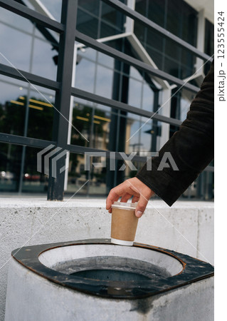 Close-up cropped shot of unrecognizable business man throwing disposable coffee cup into street trash can near office building, demonstrating responsible waste disposal and urban cleanliness. Close-up cropped shot of unrecognizable business man throwing disposable coffee cup into street trash can near office building, demonstrating responsible waste disposal and urban cleanliness. 123555424