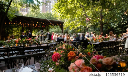 Outdoor wedding reception in lush garden setting with guests seated at long tables decorated with vibrant floral arrangements. 123556719