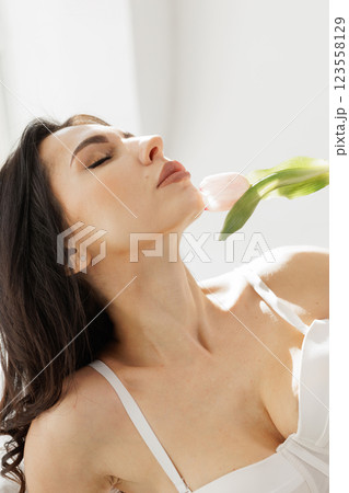 Elegant woman in white lingerie with delicate gloves holding a pink tulip near lips in soft natural light 123558129