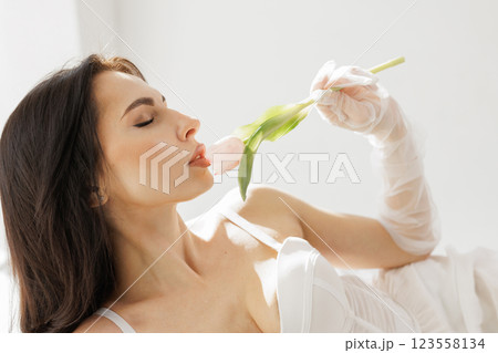 Elegant woman in white lingerie with delicate gloves holding a pink tulip near lips in soft natural light 123558134