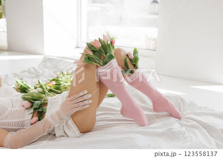 Elegant woman in lingerie lying on a bed with fresh tulips, wearing sheer gloves and pink socks, in a bright, minimalistic interior 123558137