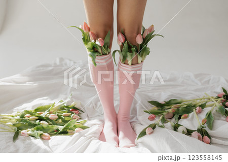 Legs in pink socks filled with fresh tulips on a white bed, playful floral fashion concept, soft natural light aesthetic. 123558145
