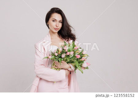 Smiling woman in a pink suit holding a bouquet of fresh tulips, posing against a neutral background in a soft, feminine, and elegant spring-inspired look 123558152