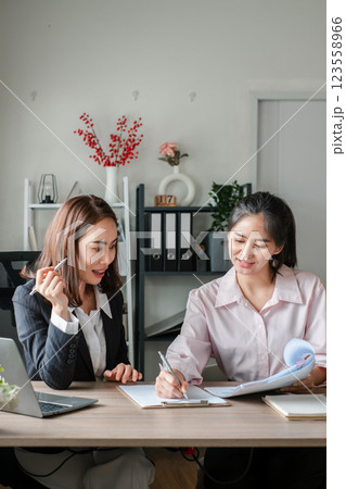 Two women in a modern office setting discussing business plans, surrounded by documents and a laptop. 123558966