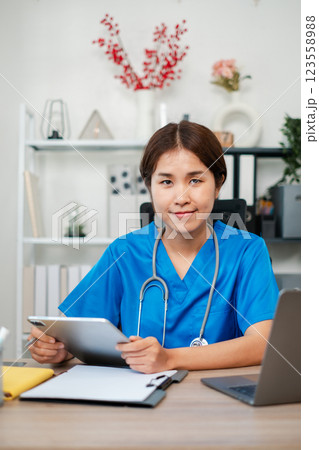 A young female nurse in blue scrubs using a digital tablet in a modern medical office setting, with a stethoscope and laptop on the desk. A young female nurse in blue scrubs using a digital tablet in a modern medical office setting, with a stethoscope and laptop on the desk. 123558988