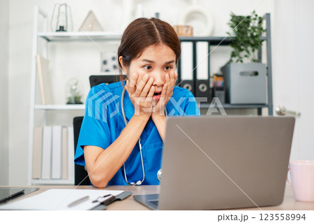 A healthcare worker in blue scrubs looks shocked while viewing a laptop in a contemporary office environment. A healthcare worker in blue scrubs looks shocked while viewing a laptop in a contemporary office environment. 123558994