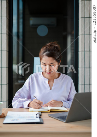 Woman sitting at a table writing in a notebook with a laptop and documents, enjoying a casual work setting. 123559007