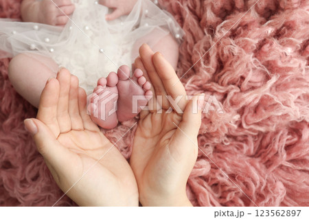 Mother holding the feet of a newborn baby on a background of pink flakatti. Feet of a newborn in the hands of parents. Macro photo of the foot, heels and toes. Openwork white dress Parents' palms. 123562897