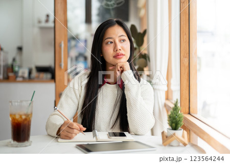 An asian woman is thinking while writing with pencil in notebook, with phone and tablet on the table 123564244