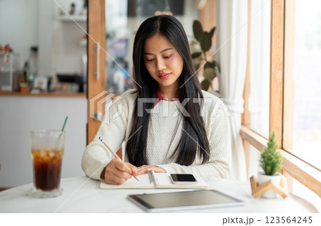 An asian woman is writing with a pencil in notebook with a smartphone and a tablet placing beside. 123564245
