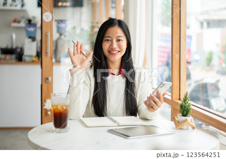 A smiling asian woman is waving to the camera while holding the phone and sitting at the cafe table. 123564251