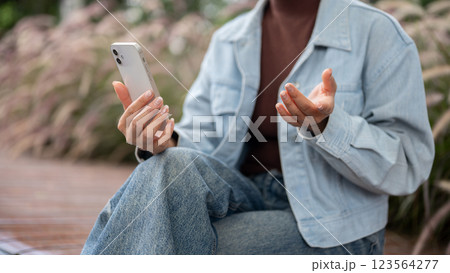A person in denim is talking to the phone while sitting on bench. Video calling or recording herself 123564277