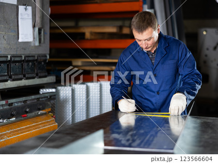 Male worker at metallurgical plant measures an iron sheet using tape measure and marks cutting line 123566604