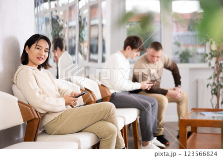 Girl sits in lobby of employment office, waiting for interview or conversation with career consultant. Watches news on Internet via smartphone Girl sits in lobby of employment office, waiting for interview or conversation with career consultant. Watches news on Internet via smartphone 123566824